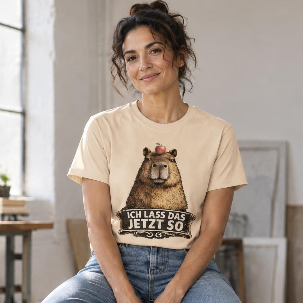 A person in a black capybara graphic t-shirt and denim jeans sitting on an industrial stool in a studio.